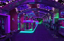 A nightclub themed as an underground subway station, with purple and green neon lights, featuring vintage subway carriages for tables and chairs. The walls have brickwork patterns and bar counter designs. A large black metal archway leads to the entrance of another club in a different part of the building.