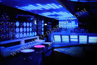 A wall of LED light tubes in nightclub in Germany. Lightning DJ booth, big sofas with three tables left from the DJ. The lights glow blue and white, creating an atmosphere that is both futuristic yet warm and inviting. In front of it stands a table displaying various products.