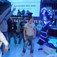 A group of young men and women(Disco Designer`s staff) at an event in Monaco were having fun dancing on the dancefloor with their hands up, smiling for the camera. The room was lit by blue LED lights and had white furniture. In front there was one DJ table with turntables and music equipment.
