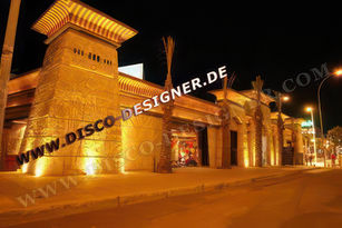 A club exterior in the style of an ancient Egyptian building, decorated with hieroglyphs and columns made from golden stone. The entrance is illuminated by warm yellow lights that reflect off its surface.