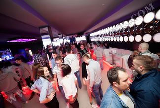 Photo of the interior of a disco club in Riga, with people dancing and drinking. The room is filled with colorful lights and large disco balls hanging from above. There is an energetic atmosphere lit with neon light and laser beams. A crowd fills up on the dancefloor enjoying music and having fun.