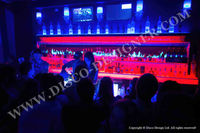 an LED bar display at the top of an elegant, modern club shelf in Miami with women dancing on it and people drinking drinks. 