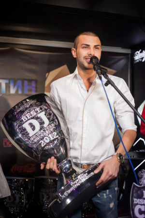 Photo of a smiling man in a leather jacket holding a large trophy cup with text "best club in Bulgaria" on it, photorealistic, on stage near the microphone under studio lighting.