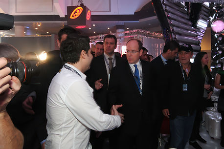 A man in a white shirt and blue tie(CEO of Disco Designer) shaking hands with the Prince of Monaco at mICS Show 2011 in Monaco, surrounded by photographers and cameras. Flashlights were flashing everywhere as people in black suits took professional photographs.
