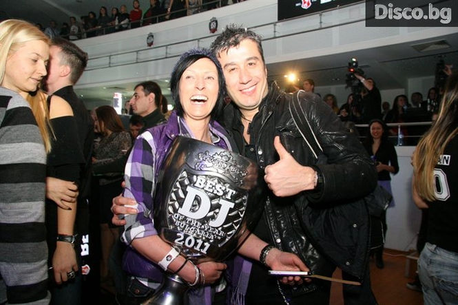 The awards ceremony of the "Best club in Bulgaria" project, which takes place in Bulgaria. A trophy with text written on it stands next to the stage where two people stand behind a LED screen displaying music on one side, while on another there is a beautiful woman dressed as Eastern European, wearing black dress made out of diamonds holding up a big award.