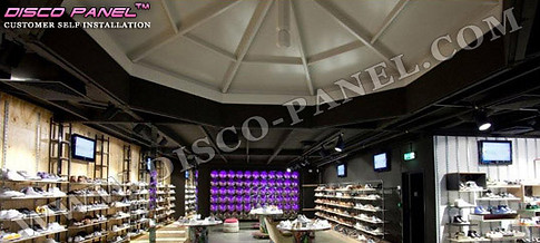 Dreadfull disco panel light installation in an open shoe store with shelves full of shoes and two flat screen televisions on the wall. The ceiling is made out of dark wood, purple led lights.