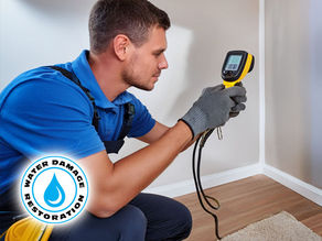 technician using a handheld thermal imaging device on a living room wall, with the screen showing a cold spot. Moisture meters and drying fans are visible nearby, creating a professional inspection setting for leak detection