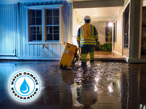 damage restoration technician in waterproof boots and gear stepping into a flooded living room at night, with drying equipment being set up—exterior shows a West Palm Beach home during a storm