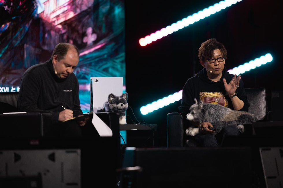 Two men sit on stage; one writes, the other gestures while holding a wolf plush toy. A PlayStation and neon lights are in the background.