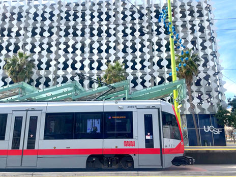 A muni train is pulling up to an outdoor train stop in Mission Bay, in front of a UCSF building with a white modern facade.