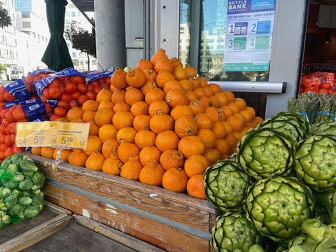Image taken of the exterior of a small grocery store, featuring a pyramid of oranges on a wooden stand, with artichokes on the right and bags of Brussels sprouts on the left.
