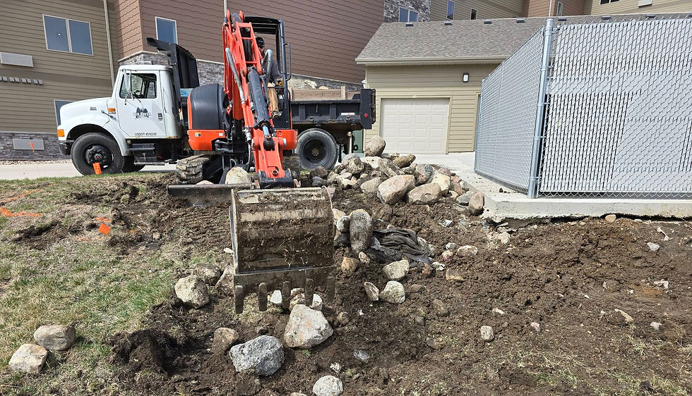 Excavator removes large rocks from a grassy area beside a chain-link fence, with a dump truck parked nearby.
