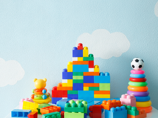 Blocs colorés en plastique empilés en forme de pyramide, ours en jouet, anneaux arc-en-ciel et ballon, sur fond bleu avec nuages blancs.