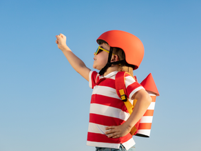 Garçon en casque orange et lunettes, bras levé, avec sac à dos, sur fond de ciel bleu.