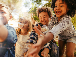 Quatre enfants rient et attrapent des bulles dans un parc ensoleillé, sous les arbres, ambiance joyeuse.