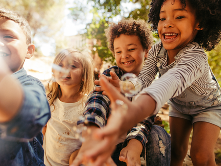 Quatre enfants rient et attrapent des bulles dans un parc ensoleillé, sous les arbres, ambiance joyeuse.