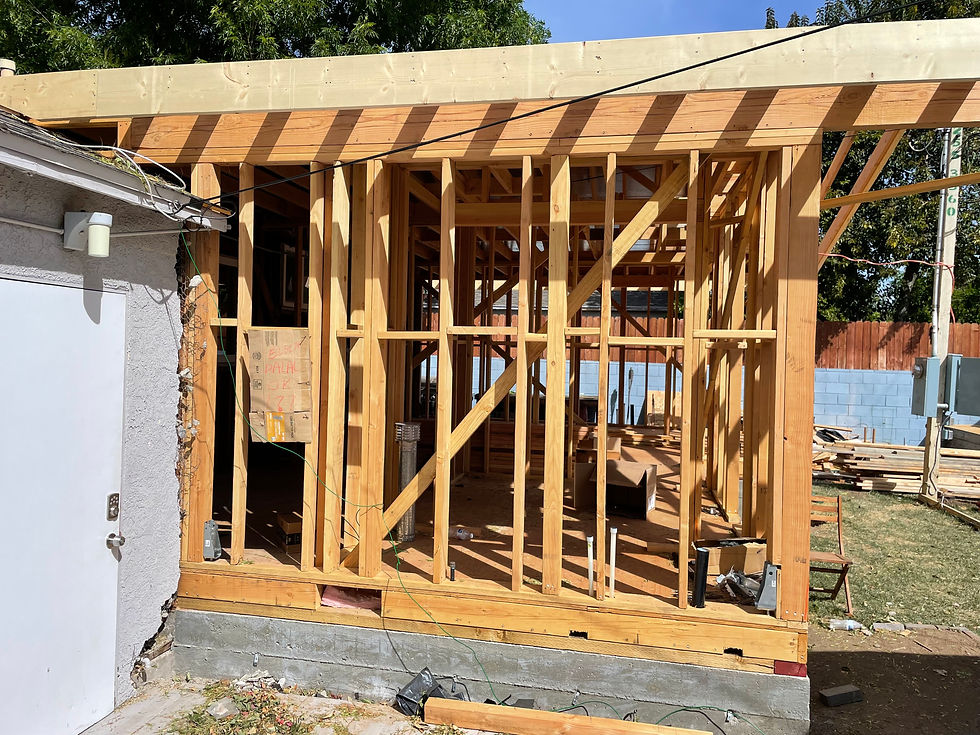 Wooden framework of a house under construction with visible yard and trees. A cardboard sign with handwriting hangs inside. Bright daytime setting.