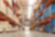 A wide-angle view of a large warehouse aisle, with tall shelves on both sides stocked with various boxes and containers, extending into the distance