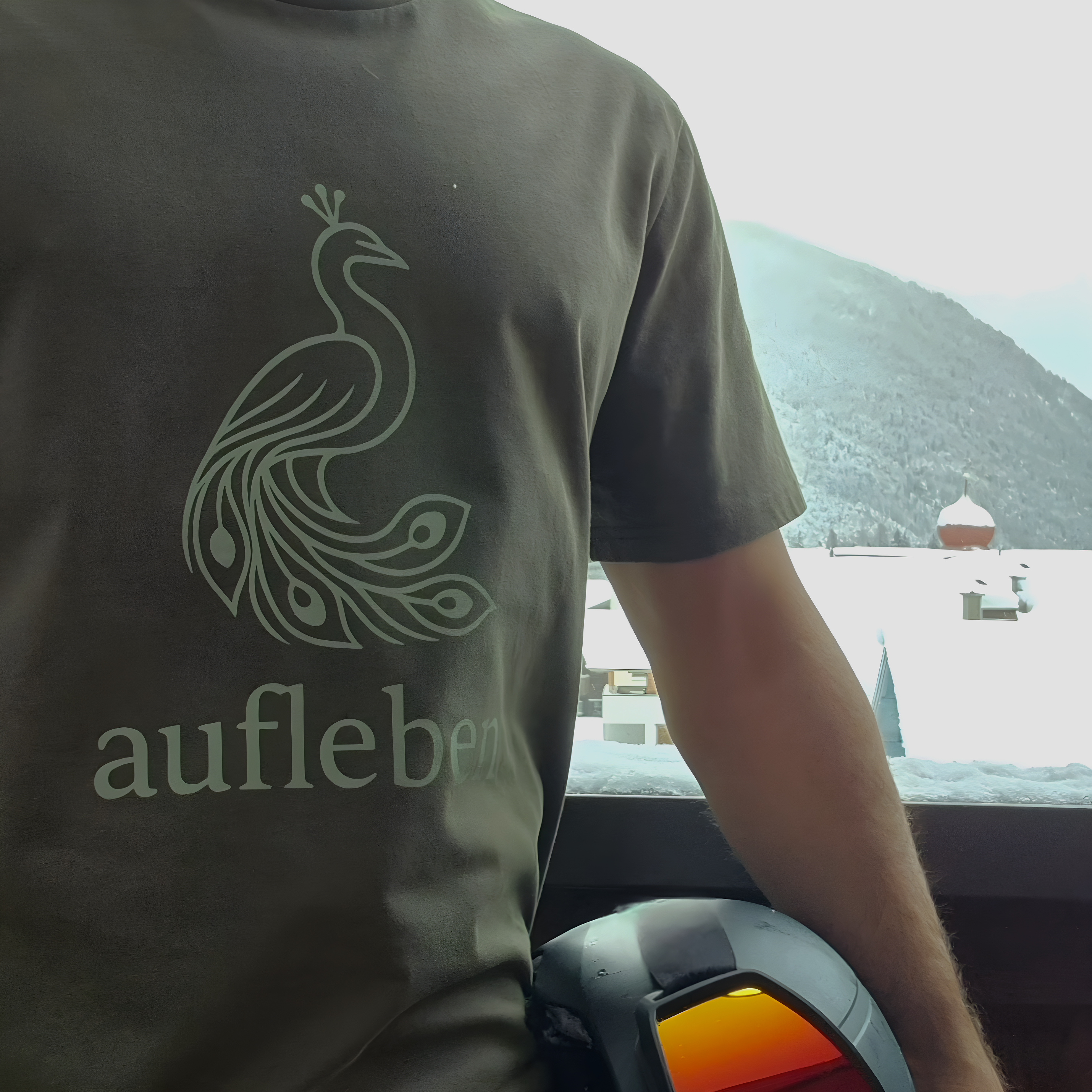 A person wearing an olive green aufleben T-shirt with a peacock design, standing on a balcony overlooking a snowy mountain la