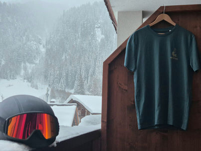 Organic deep green t-shirt by aufleben with small peacock chest logo, hanging on wooden balcony in winter mountain backdrop