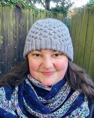 Su faces the camera. A white woman with rosy cheeks and brown hair wearing a grey knitted hat and a blue striped knitted shawl.