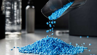 Blue plastic pellets being dropped out of a metal scoop onto a table top.
