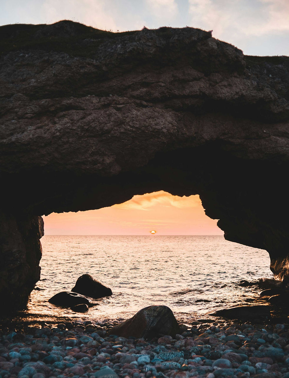 Rocky beach with a stone arch framing a sunset over the sea. Warm orange hues dominate the sky, creating a serene and natural mood.