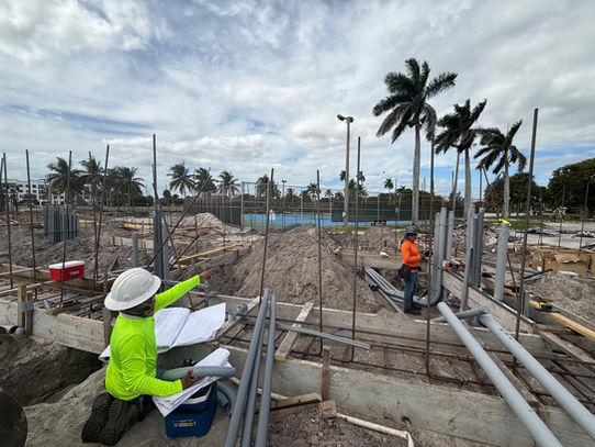 Construction workers review blueprints at a building site, palm trees in the background.