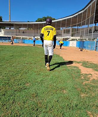 Dominican high school baseball player in yellow uniform walking to bat next, on deck