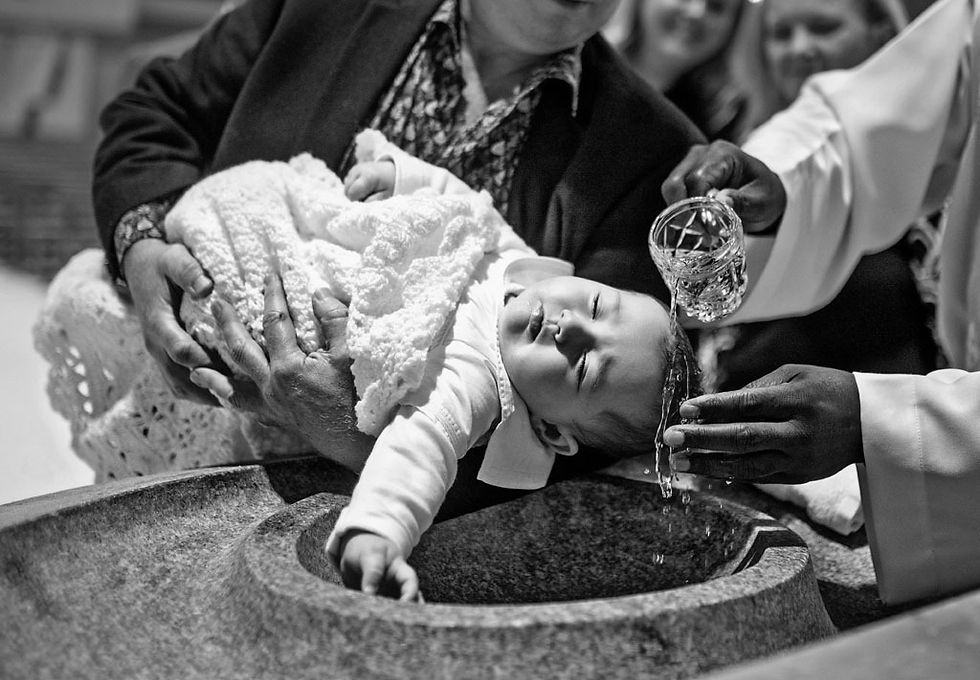 baby boy wrapped in white blanket having head wetted at baptismal font in church as dad holds him. showing priest pouring water on baby's head. emotive baptism photographer dublin. bapism family photoshoot, in church and celebration after