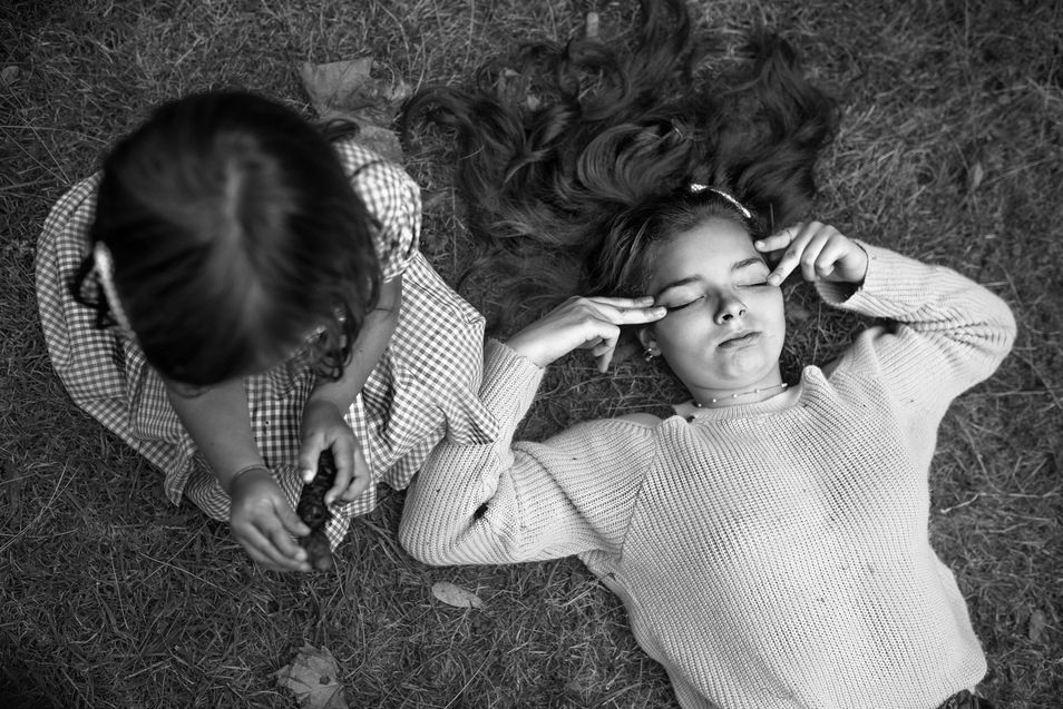 little sister and big sister lying out on the grass with big sister covering her eyes with her hands to avoid glare from sun