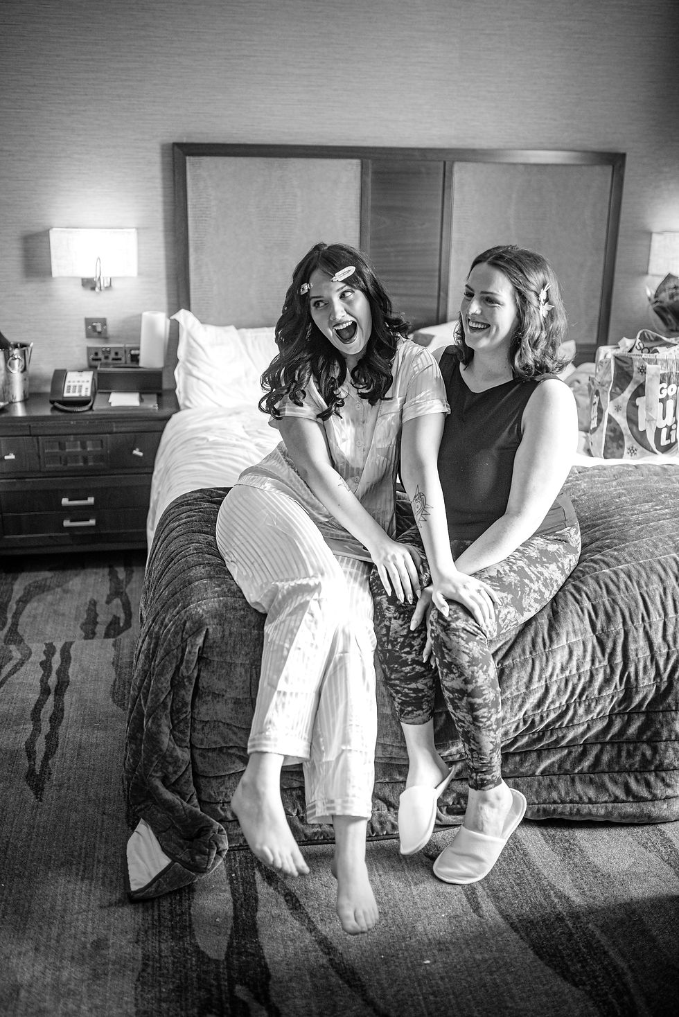 bride and bridesmaid laughing on bed in PJ's and slippers and with giant hair clips
