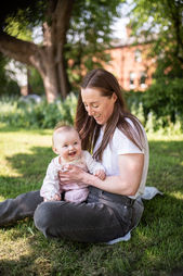 young woman in jeans and T shirt sitting in softly sunlit park with baby girl in pink