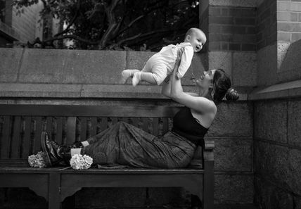 mum holding baby boy high over her head as she lies back on a bench in a garden courtyard