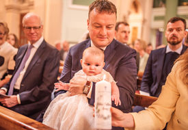 dad in suit holding baby in the pews of church as mum holds her christening candle