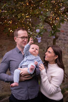 dad holding baby boy with mum as he reaches for fairy Christmas lights hanging from tree
