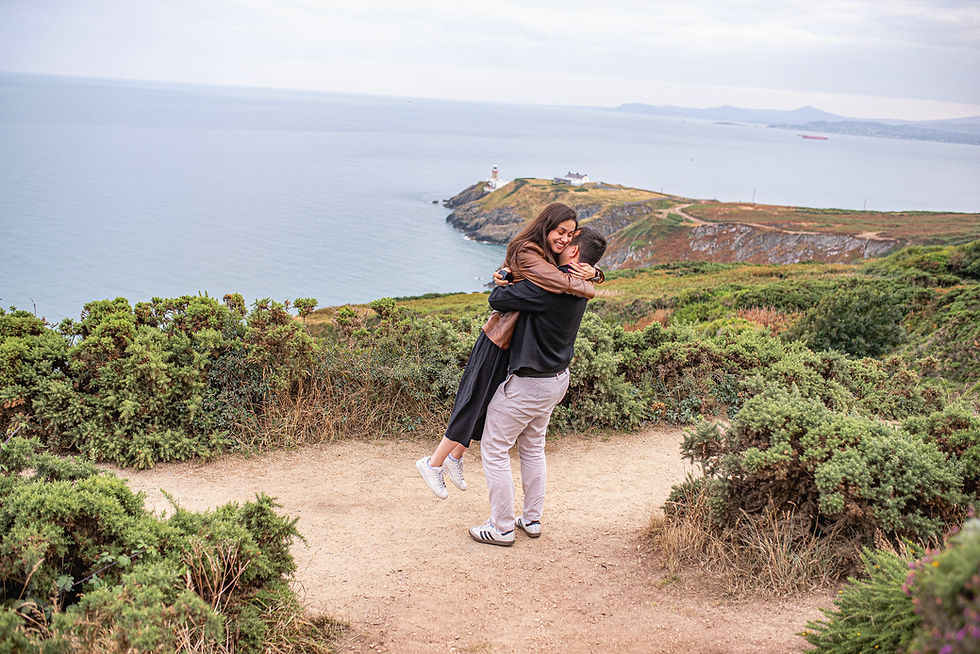 young woman in brown leather jacket jumping into her boyfriend's arms on Howth cliff top opposite the lighthouse with view of the ocean and heather