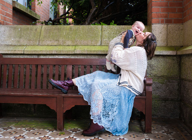mum in vintage flowing blue skirts and baggy cardigan lifting baby boy on a bench in a court yard and kissing his cheeks