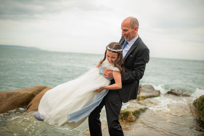 dad in suit twirling his daughter around in a communion dress with sea in the background