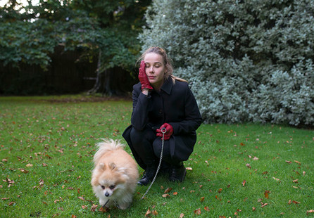 young oman holding hand in red leather glove to her face with dog on a lead. she has her eyes closed. background of autumn leaves and a blue bush