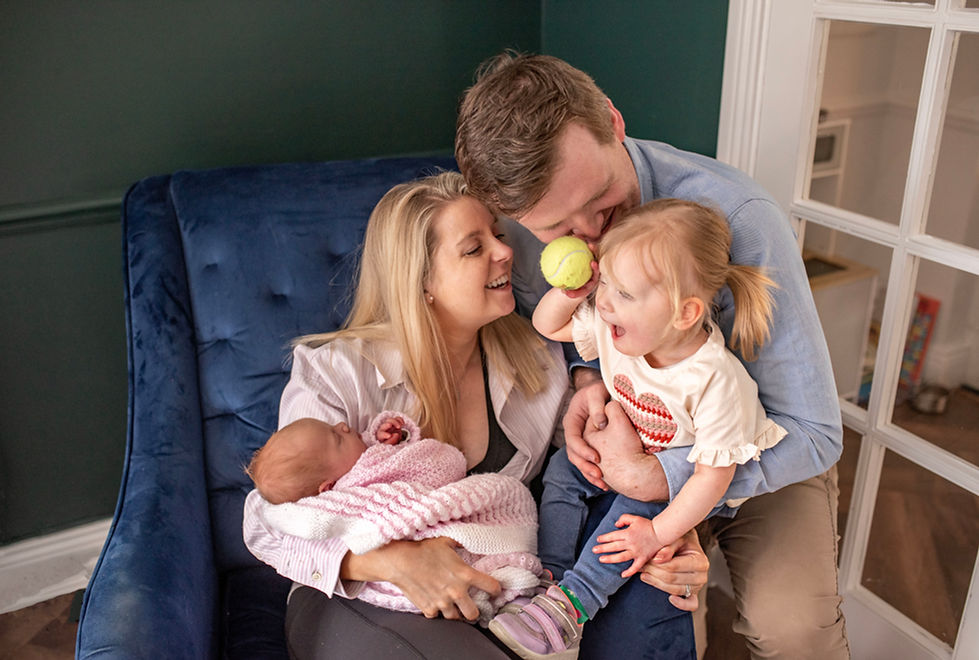 mum, dad on armchair with newborn baby girl and older sister holding a tennis ball