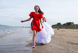 girl in communion gown running along beach with older sister in red dress