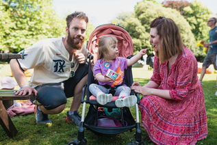 mum and dad either side of a pram with toddler in pram leaning towards mummy and spoon feeding her yoghurt from a tub