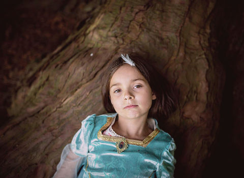 girl in a princess costume with a white feather in her hair lying against a tree trunk