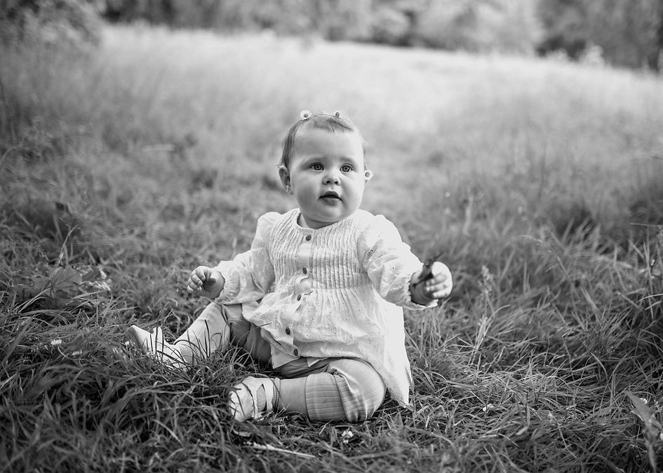 baby girl sitting in a meadow in white cotton dress. daisies in her hair and holding leaf. spring baby photo shoot dublin.