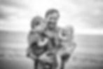 dad holding his two young sons with baby laughing at the beach