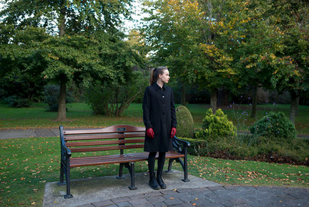 woman standing gracefully in dark coat and red gloves next to a park bench in avoca park, blackrock dublin