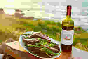 A wooden table with a place of freshly cooked food and a bottle of St Martin's wine on a clifftop with the sea behind