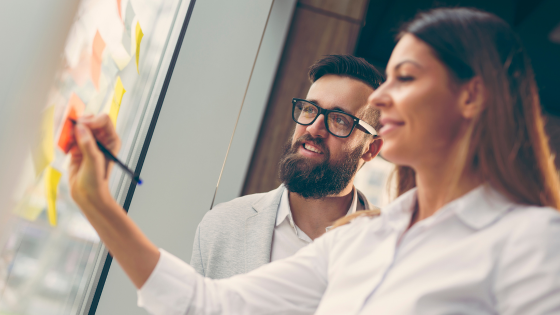 Two business professionals developing a supply chain analytics strategy on a glass board with post it notes.