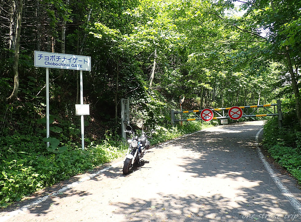 森の中の道路で、チョボチナイゲートの看板と通行止め柵が見え、手前にバイクが停まっている。